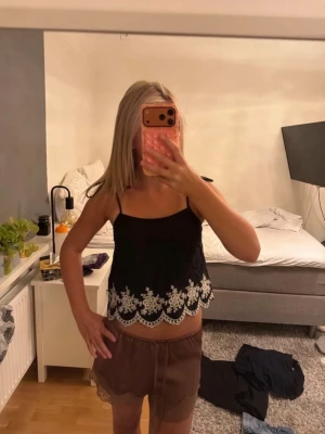 Svart linnetopp med spets från H&M - Säljer en svart linnetopp från H&M Divided med tunna axelband och vit spetsdetalj längs nederkanten. Toppen är croppad och har ett romantiskt broderat mönster som ger en söt vibe. Perfekt till sommaren och enkel att matcha med jeans eller kjol.