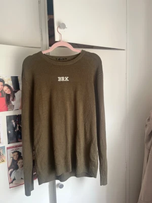 Olivgrön stickad tröja BRK - Olivgrön stickad tröja med texten 'BRK' framtill och 'BROOKLYN 82' i vitt på ryggen. Tröjan har rund halsringning och ribbade muddar vid ärmslut och nederkant. Perfekt för en avslappnad streetstyle-look.