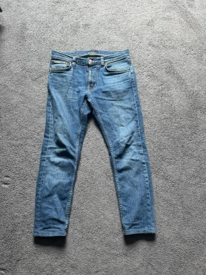 Blå jeans med smal passform - Säljer ett par klassiska blå jeans från Nudie Jeans med smal passform. W29 L28 Jeansen har fem fickor, orangea sömmar och Nudie-loggan på bakfickan. Tillverkade i denim med snygg tvätt och subtila slitningar. Perfekta för en avslappnad och trendig stil. 