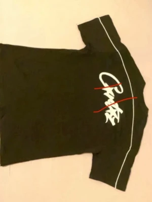 Svart t-shirt med vit text och linjer - Svart t-shirt med vita tryck och grafiska detaljer på både fram- och baksidan. T-shirten har korta ärmar och vita linjer längs axlarna. Materialet ser ut att vara bomull, vilket gör den skön att bära. Perfekt för en avslappnad och trendig stil.