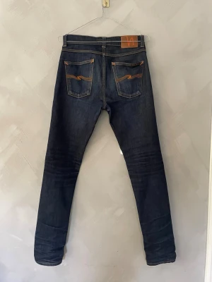 Mörkblå nudie jeans - lean dean dry 16 dips - Säljer ett par mörkblå jeans från Nudie Jeans med klassisk femficksmodell och kontrastsömmar. Jeansen har raka ben och läderpatch bak i midjan. Modellen är lean dean och det är torr denim vilket gör att jeansen bygger sjukt snygga fades med tiden. Det har redan påbörjats några fades men med mer tid kan de bli sjukt snygga.