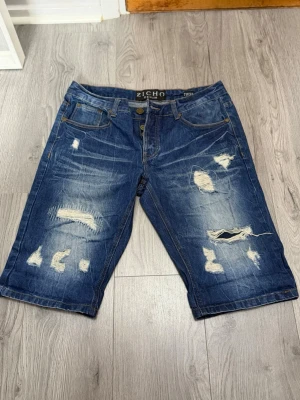 Blå slitna jeansshorts från Zicho - Säljer ett par blå jeansshorts från Zicho med flera slitningar och trasiga detaljer både fram och bak med knappgylf och råa kanter