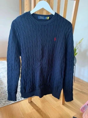 Mörkblå kabelstickad tröja Polo Ralph Lauren - Kabelstickad mörkblå tröja från Polo Ralph Lauren med rund halsringning och röd broderad logga på bröstet. Tröjan har ribbade muddar vid ärmslut och nederkant och är tillverkad i mjukt material. Perfekt för en stilren och klassisk look. Obs! Kan ej garantera äktighet då den köpt på vinted därav priset!