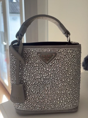 Grå glittrig handväska från Prada - Superfin grå handväska från Prada täckt med massor av glittrande stenar. Väskan har en stadig form, kort handtag och detaljer i silver. Insidan är rymlig med flera fack. Perfekt för dig som vill sticka ut med en lyxig accessoar. Helt oanvänd. 