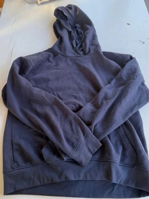 Mörkblå basic hoodie med huva - En enkel och stilren mörkblå hoodie med huva och känguruficka framtill. Hoodien har ribbade muddar vid ärmslut och nederkant. Perfekt för en avslappnad och chill look. Materialet känns mjukt och bekvämt, troligtvis bomullsblandning.