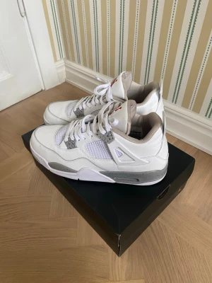 Vita Air Jordan 4 sneakers - Snygga vita Air Jordan 4 sneakers med grå och silverdetaljer. Skorna har meshpaneler på sidorna, klassisk snörning och Jumpman-logga på hälen. Ovandelen är i läder och mesh, och sulan är vit med grå inslag. Perfekta för dig som gillar streetwear.