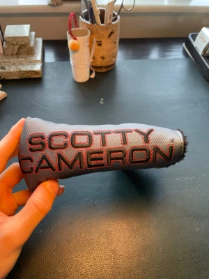 Scotty Cameron putter headcover grå - Säljer ett grått putter headcover från Scotty Cameron med broderad röd text och guldfärgad krona på sidan. Skyddet är tillverkat i slitstarkt tygmaterial och har en klassisk L-form som passar de flesta putters. Perfekt för golfare som vill ha stil på utrustningen. och en scotty camron green lagare