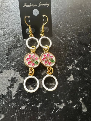 Hängande örhängen med blommor och ringar - Säljer ett par unika hängande örhängen med guldfärgade detaljer, vita ringar och en rund mittdel med rosa blomma och gröna blad. Blommans mitt är dekorerad med små pärlliknande stenar som ger extra bling. Perfekt för dig som vill sticka ut med färg och form!