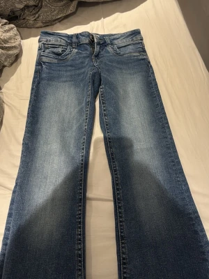 Blå bootcut jeans  - Säljer ett par klassiska blå bootcut jeans med snygga slitningar och ljus tvätt. Jeansen har fem fickor, bälteshällor och knappgylf. Perfekta för en avslappnad och trendig look.