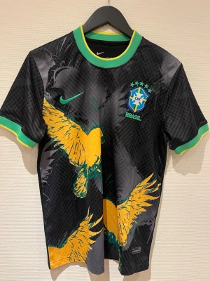 Svart Brasilien fotbollströja Nike - Snygg svart fotbollströja från Nike med Brasilien-emblem och gröna detaljer. Tröjan har ett stort gult fågelmotiv framtill, gröna och gula muddar samt rund halsringning. Perfekt för dig som vill sticka ut på planen eller heja på Brasilien.