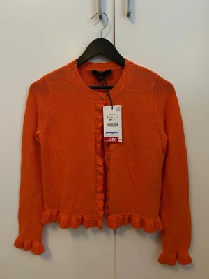 Orange kofta med volangdetaljer från Zara - Snygg orange kofta med volangkant vid ärmslut och nederkant. Rund halsringning och knappar framtill. Perfekt för dig som vill ha en färgklick i garderoben och gillar feminina detaljer. Stickad i mjukt material och har lång ärm.