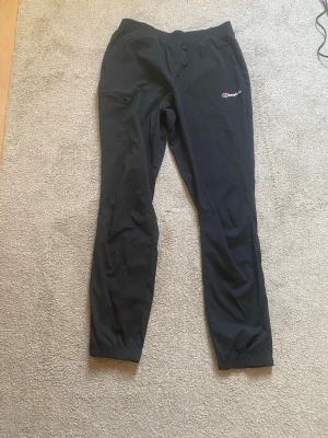 Svarta mjukisbyxor från Berghaus - Svarta mjukisbyxor från Berghaus med resår i midjan och vid bensluten. Byxorna har en enkel, sportig design med en liten logga på ena benet och en dragkedjeficka på sidan. 