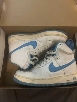 Vita och blå Nike Air Force 1 High - Säljer ett par klassiska Nike Air Force 1 High sneakers i vitt med ljusblå detaljer och sula. Skorna har högt skaft och är lite skitig men annats helt oskadd