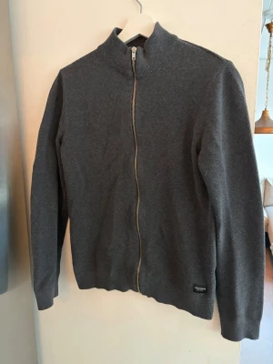 Mörkgrå ziptröja från Jack & Jones - Mörkgrå ziptröja från Jack & Jones med hög krage och hel dragkedja framtill. Tröjan är stickad i mjukt material och har ribbade muddar vid ärmslut och nederkant. Perfekt för lager på lager och enkel att matcha med olika outfits. Storlek xs passar S