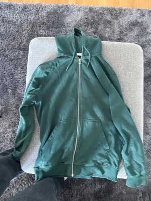 Grön hoodie med dragkedja från Weekday - Mysig grön hoodie från Weekday i storlek XS. Hoodien har dragkedja framtill, fickor på magen och justerbar huva med snören. Perfekt för en chill och avslappnad stil. Tillverkad i mjuk bomull.