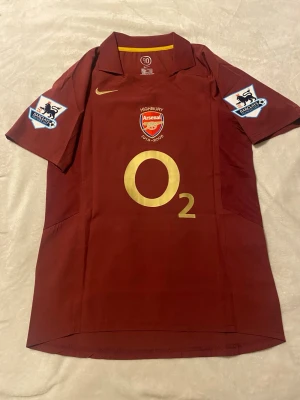 Arsenal Highbury tröja Henry 14 - Säljer en vinröd Arsenal Highbury-matchtröja från Nike med korta ärmar, guldfärgade detaljer och tryck. Tröjan har klubbmärke, O2-logga och Premier League-patchar på ärmarna. På ryggen står det Henry 14 i guld och Highbury 1913-2006. Helt ny. Pris kan sänkas vid snabb affär