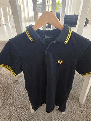 Svart Fred Perry piké med gula detaljer - Klassisk svart pikétröja från Fred Perry med gula ränder på krage och ärmslut samt den ikoniska loggan broderad på bröstet. Tillverkad i bomull och har normal passform med korta ärmar och knappar vid halsen.