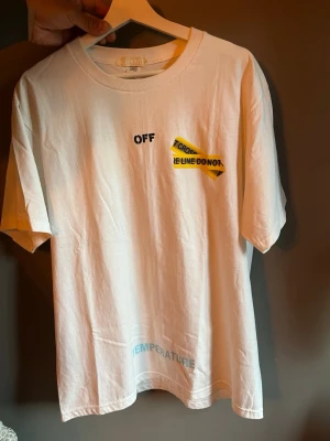 Vit Off-White T-shirt med tryck - Säljer en vit T-shirt från Off-White med svart text och gul tejpdetalj på bröstet samt stort grafiskt tryck med 'FIRE LINE DO NOT CROSS' på ryggen. T-shirten har rund hals och korta ärmar. Ordet 'TEMPERATURE' är tryckt i ljusblått nedtill.