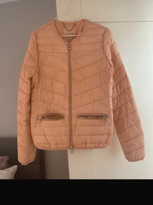Ljusrosa pufferjacka från Amisu - Snygg ljusrosa pufferjacka från Amisu med raka sömmar och två dragkedjeförsedda fickor framtill. Jackan har rund halsringning och dragkedja hela vägen fram. Tillverkad i ett lätt, quiltat material som håller värmen under kalla dagar.