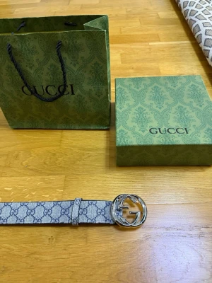 Gucci bälte med silverfärgat spänne - Säljer ett snyggt Gucci bälte med klassiskt GG-mönster i grått och svart. Bältet har ett stort, rundat spänne i silver och levereras med originalpåse och ask. Perfekt accessoar för att lyfta din outfit.