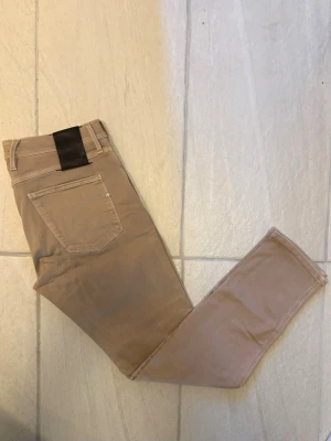 Replay jeans herr beige  - Snygga beige jeans med rak passform och klassisk femficksdesign. Jeansen har bälteshällor och en diskret detalj på bakfickan. Perfekta för en clean och stilren look.