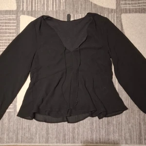 Svart blus med v-ringning från Divided - Svart blus från Divided H&M med v-ringning och tunna knytband framtill. Blusen har långärmad design och är tillverkad i ett lätt, något transparent tyg som ger ett flowy intryck. Perfekt att styla med jeans eller kjol.