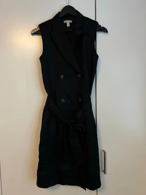 Svart ärmlös kortklänning med bälte - Snygg svart ärmlös kortklänning med dubbelknäppning framtill och matchande bälte i midjan. Klänningen har krage och en stilren, klassisk look som passar perfekt till många tillfällen. Enkel att styla med accessoarer för en personlig touch.