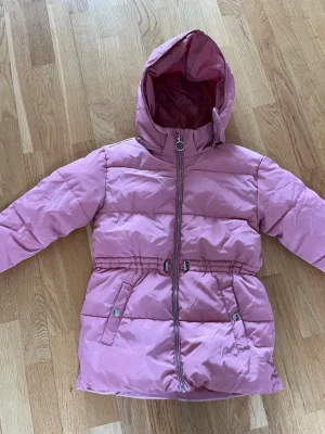 Rosa pufferjacka från KappAhl - Snygg rosa pufferjacka från KappAhl med huva och dragkedja framtill. Jackan har två fickor med tryckknappar och elastisk midja för skön passform. Perfekt för kalla vinterdagar och riktigt mysig att bära. Det står den är i stl 146 men skulle säkert även passa någon med stl XXS. Den är använd men i bra skick, inga stora defekter. Hör av er om frågor!💗(Säljer även på Vinted ifall man skulle vilja köpa där)