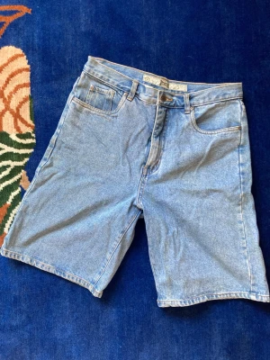 Blå jeansshorts från Guess - Klassiska blå jeansshorts från Guess med fem fickor, normal passform och dragkedja. Tillverkade i bomull med ljus tvätt och detaljerade sömmar på bakfickorna. Perfekta för sommardagar och enkel att matcha med olika stilar.