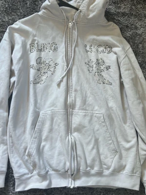 Vit hoodie från Blinglucid med bling - Säljer en vit hoodie från Blinglucid med dragkedja, huva och fickor framtill. På bröstet finns glittriga detaljer och texten 'BLING LUCID' i stenar samt två figurer. Hoodien är i mjukt material och har en avslappnad passform.och sedan så har den gått sönder god fikan en gång men det är igen sytt och sina knapt.