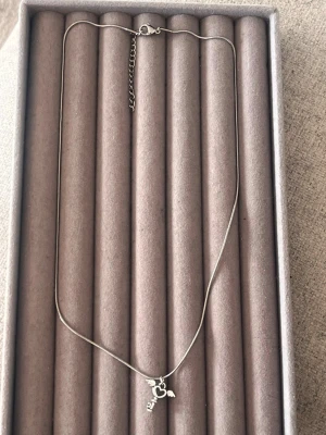 Silverfärgat halsband med hjärthänge - Säljer ett tunt silverfärgat halsband med ett hänge i form av ett hjärta med små vingar och en nyckel. Kedjan är enkel och smal, och hänget ger en söt och lekfull touch. Perfekt för dig som gillar detaljerade smycken med symbolik.