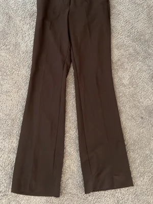 Bruna vida kostymbyxor - Snygga bruna kostymbyxor med vida ben och klassisk passform. Byxorna har hög midja och är tillverkade i ett mjukt, vävt tyg som ger en stilren look. Perfekta för dig som gillar en clean och trendig stil.