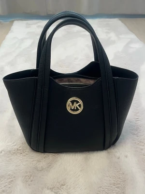 Svart handväska Michael Kors - Snygg svart handväska från Michael Kors i stilren design med guldfärgad MK-logga framtill. Väskan har två handtag och är gjord i ett tåligt, strukturerat material som ger en lyxig känsla. Perfekt storlek för det viktigaste och enkel att matcha till olika outfits.