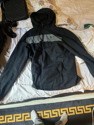 Svart vindjacka från Under Armour - Snygg svart vindjacka från Under Armour med grått band och logga över bröstet. Jackan har huva med dragsko, hel dragkedja framtill och två sidofickor. Tillverkad i lätt och vindtätt material, perfekt för blåsiga dagar.
