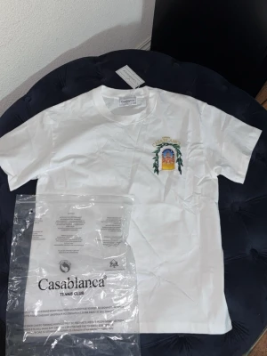 Vit Casablanca t-shirt med tryck - Säljer en fräsch vit t-shirt från Casablanca med färgglatt tryck på bröstet och diskret logga. T-shirten har klassisk passform och är gjord i mjuk bomull, perfekt för en clean och stilren look.