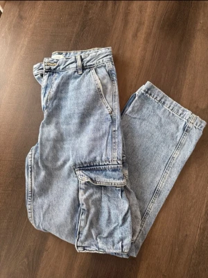 Ljusblå cargobyxor i denim jeans - I fint skick. Säljer ett par ljusblå cargobyxor i denim med raka ben och stora fickor på sidorna. Klassisk femficksmodell med bälteshällor och dragkedjegylf. 