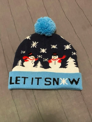 Blå mössa med snögubbar och tofs - Mysig blå mössa med ljusblå uppvikt kant och stor tofs på toppen. Motiv med snögubbar, granar och snöflingor samt texten 'LET IT SNOW' framtill. Perfekt för vintern och julen. Stickad i mjukt material som håller dig varm. Den har lite fläckar men syns inte mycket tycker jag. När batteriet är slut så kan man byta och lägga till en ny.