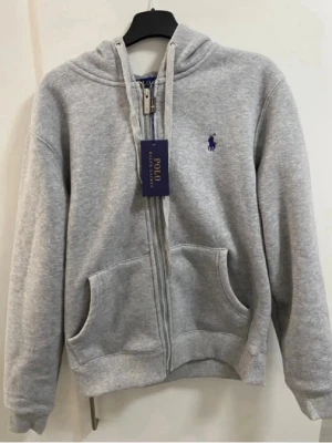 Grå zip hoodie från Polo Ralph Lauren - Klassisk grå hoodie med dragkedja från Polo Ralph Lauren. Hoodien har två stora fickor framtill, justerbar huva med snören och den ikoniska lilla loggan broderad i marinblått på bröstet. Tillverkad i mjukt sweatshirtmaterial, perfekt för en avslappnad stil. Helt ny och oanvänd