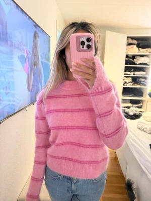 Rosa stickad mohair/wool blend med ränder - Mysig stickad tröja i ljusrosa med mörkare rosa horisontella ränder. Tröjan har rund halsringning, långa ärmar och en avslappnad passform. Perfekt för att addera färg till din garderob och passar till jeans eller kjol.k
