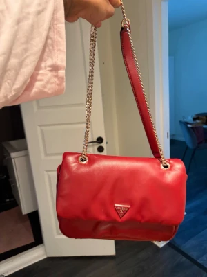 Röd axelväska från Guess - Snygg röd axelväska från Guess i mjukt konstläder. Väskan har guldfärgade kedjedetaljer, justerbar axelrem och en ficka med dragkedja på baksidan. Triangelformad Guess-logga framtill ger en trendig touch.