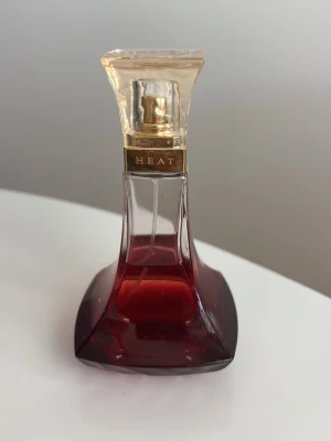 Beyoncé Heat parfym röd glasflaska - Snygg parfymflaska från Beyoncé Heat med elegant röd glasdesign och guldfärgad detalj vid halsen. Flaskan har en fyrkantig botten och smalnar av mot toppen med ett transparent lock. Perfekt för dig som gillar lyxiga och sensuella dofter. Ca 35 ml kvar i flaskan 
