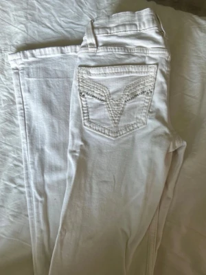 Vita jeans med broderad bakficka - Säljer ett par vita jeans med bootcut stil och snygg broderad detalj på bakfickorna. Jeansen har low waist midja och dekorativa nitar på fickorna. Skriv gärna för mina bilder på byxorna om inteserad!!  Dem passar inte mig längre men endast haft dem i 3 månader. Har även sytt upp dem i benen för dem var för långa. Jag är 1.63