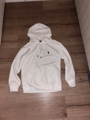 Vit hoodie med broderad logga - Säljer en clean vit hoodie med huva och dragsnören. På bröstet finns en liten broderad logga i svart. Hoodien har en stor magficka och ribbade muddar vid ärmar och midja. Perfekt för dig som gillar enkel och stilren streetwear.