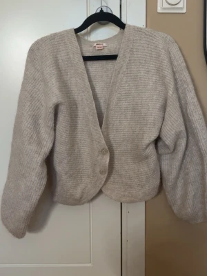 Beige stickad kofta från Gina Tricot - Mysig beige stickad kofta från Gina Tricot med två knappar framtill och avslappnad passform. Koftan har v-ringning, långa ärmar och ribbade detaljer vid ärmslut och nederkant. Perfekt för lager på lager och enkel att matcha.