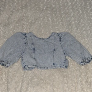 Ljusblå jeansblus från Gina Tricot - Säljer en ljusblå jeansblus från Gina Tricot med puffiga ärmar och knytning i ryggen. Blusen är croppad och har en avslappnad passform. Perfekt för en trendig look!💗💞