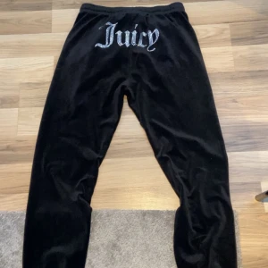 Svarta mjukisbyxor från Juicy Couture - Snygga svarta mjukisbyxor från Juicy Couture i mjukt material. Byxorna har en elastisk midja med snörning och en glittrig Juicy-logga på baksidan. Perfekta för en avslappnad stil. En av snörrenas metal sak hae gått av men annars är dom i fint skick.