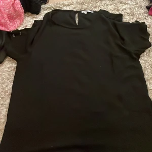 Svart blus från Flash Woman - Säljer en elegant svart blus från Flash Woman. Blusen har korta ärmar och en liten öppning vid halsen som ger en stilren look. Perfekt för en chic och enkel stil.