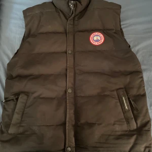 Svart dunväst från Canada Goose - Säljer en svart dunväst från Canada Goose med deras ikoniska märke på bröstet. Västen har en dragkedja och knappar framtill samt två praktiska fickor. Perfekt för kyliga dagar. Storlek XL