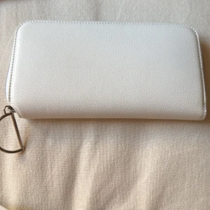 Vit plånbok från Acne Studios - Säljer en stilren vit plånbok från Acne Studios. Plånboken har en praktisk dragkedja och flera fack för kort och kontanter. Den är tillverkad i ett syntetmaterial och har en elegant metallring som detalj.