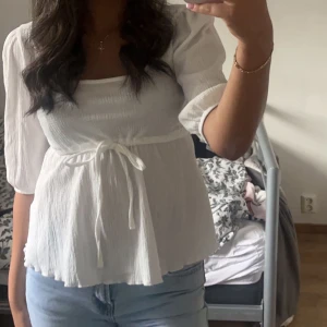 Vit blus med knytband - Säljer en söt vit blus med knytband i midjan och trekvartsärmar. Blusen har en luftig och lätt design, perfekt för varmare dagar. Den har en rundad hals och en fin struktur i tyget.